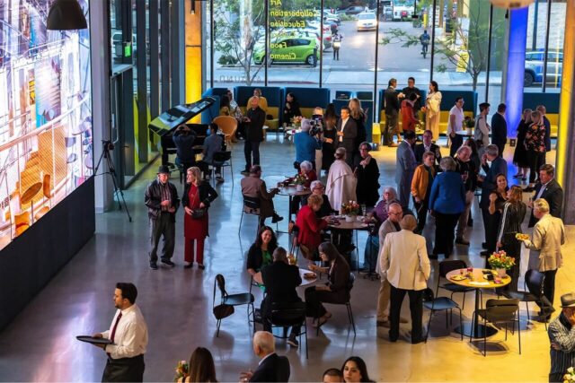 An evening exterior shot of the multi-story Park & Market building, showcasing its sleek glass and steel architecture with warm interior lights glowing through the windows and a bright digital screen visible from the street.