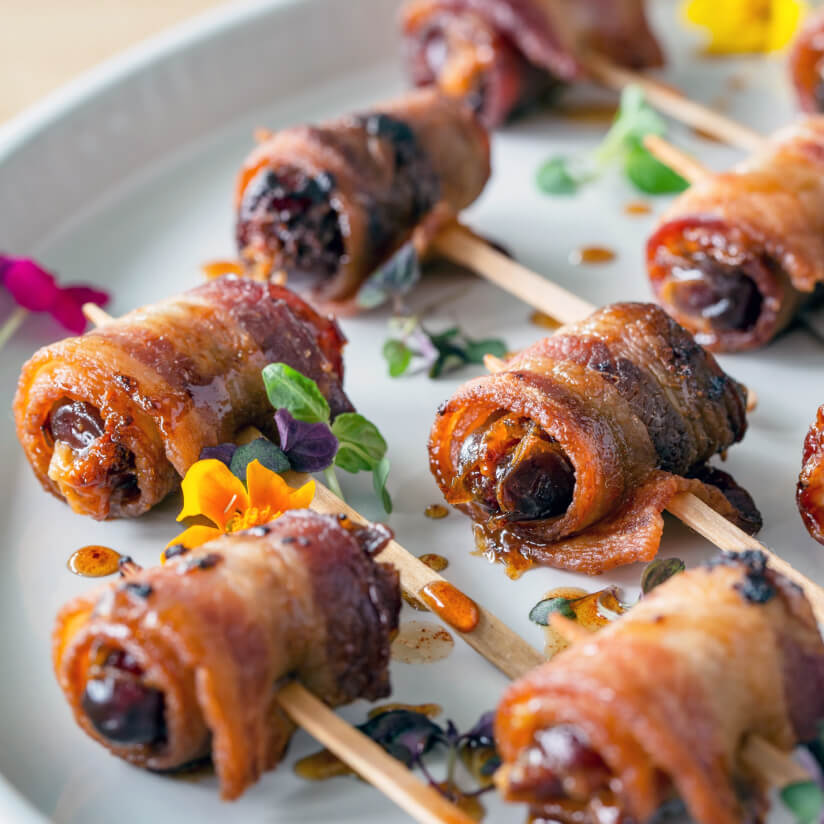 Bacon-wrapped skewers with edible flowers on a dark surface