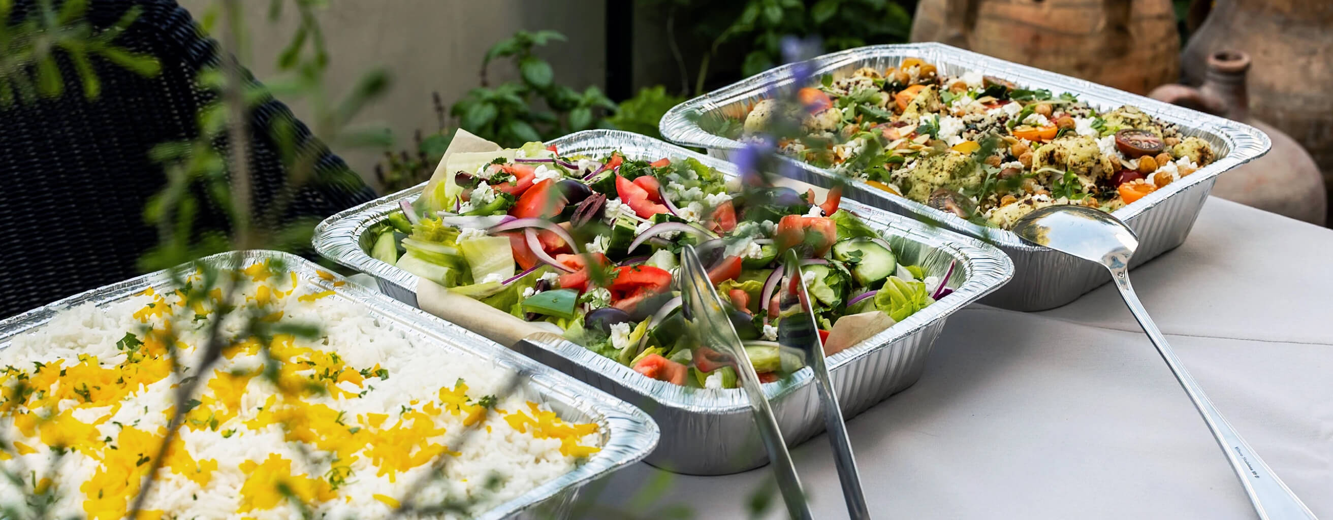 Freshly prepared catering trays of Greek salad and saffron-garnished rice served outdoors on a white linen table.