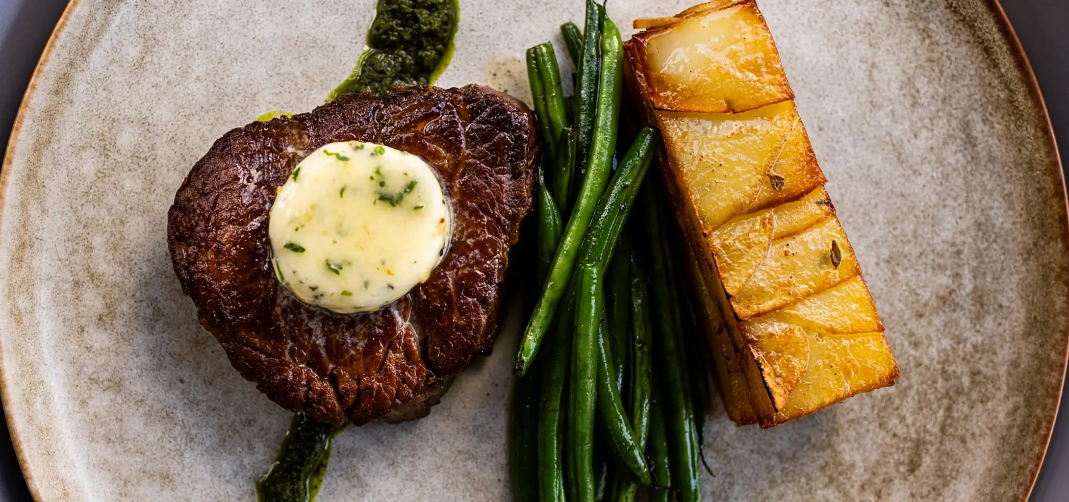 Seared steak topped with herb butter, served with green beans and roasted potatoes on a ceramic plate.