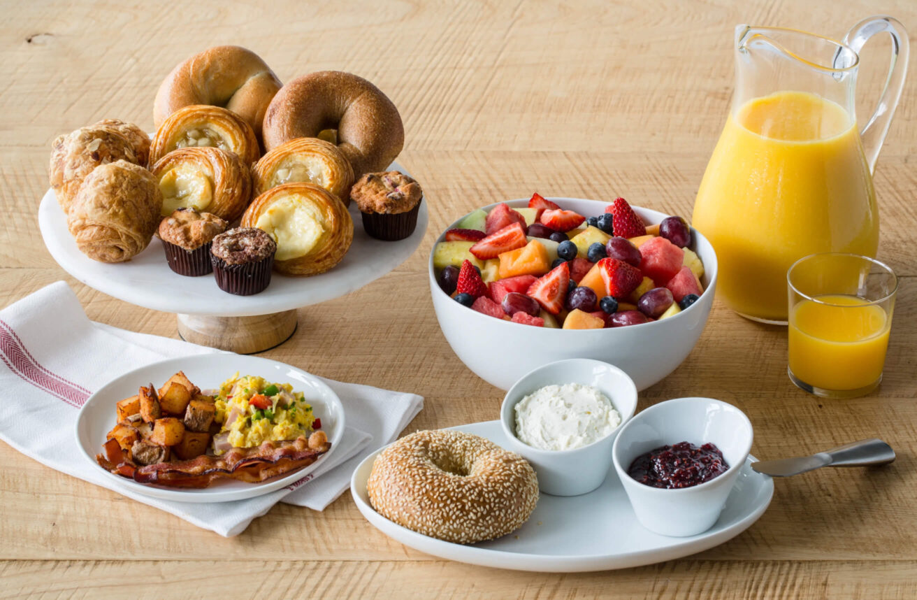 Breakfast catering spread with pastries, bagels, fresh fruit, scrambled eggs, bacon, breakfast potatoes, cream cheese, jam, and orange juice.