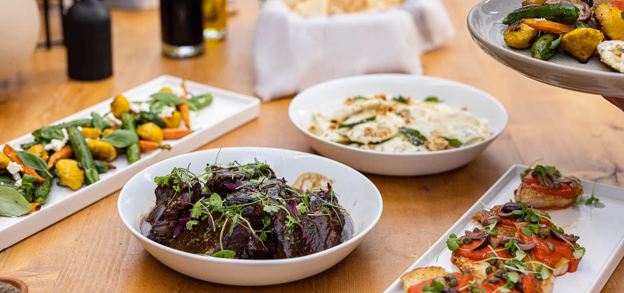 Family-style catering dishes being served at a dining table, including braised beef, roasted vegetables, pasta, and crostini prepared with fresh ingredients.