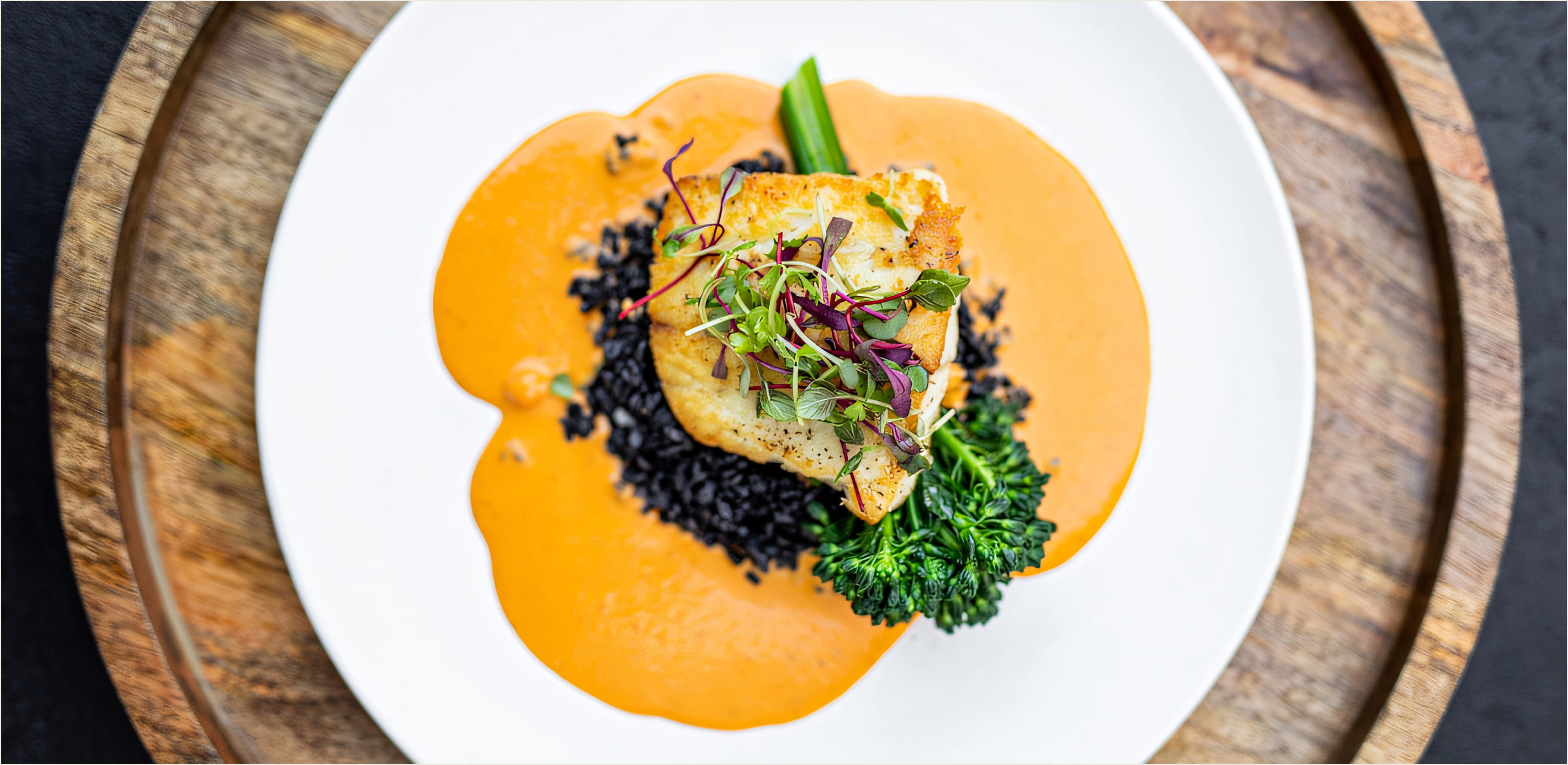 Pan-seared white fish over black rice with a swoosh of orange bisque, topped with microgreens and served with broccolini on a white plate.